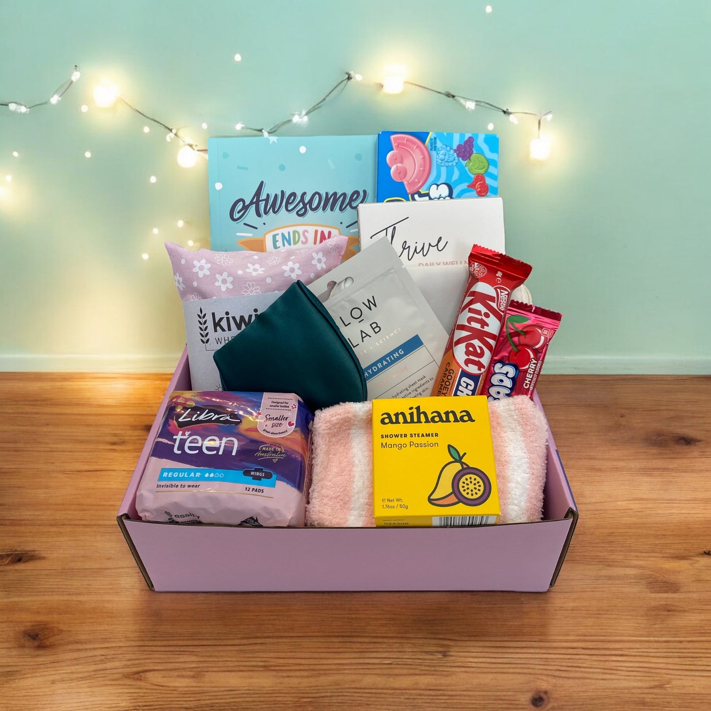 Items of gift box on disply in gift box. Awesome ends in me journal, american candy, thrive wellness cards, kiwi wheatbags, glow lab face mask, kit kat, airheads, eye mask, libra teen pads, fluffy socks and anihana shower steamer.