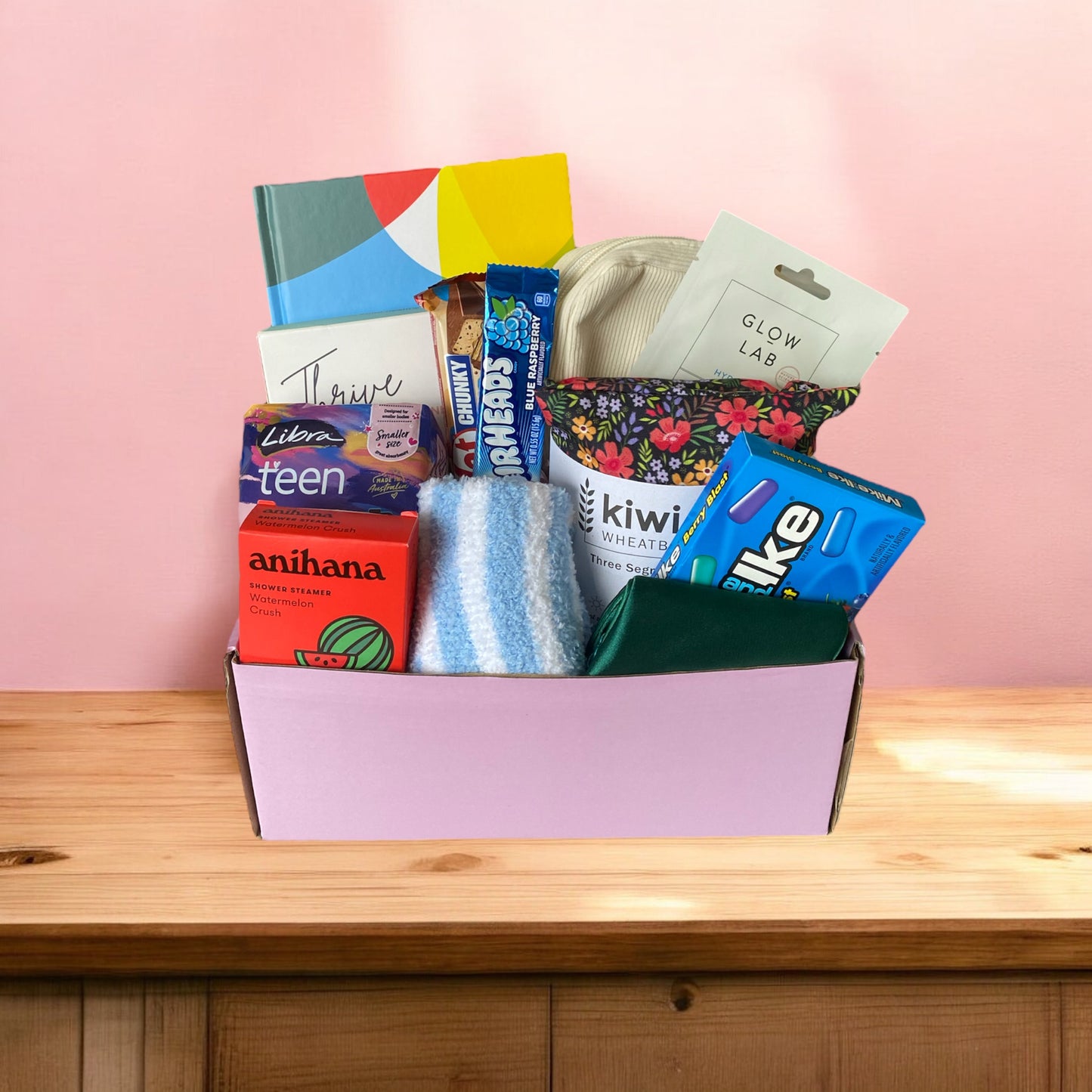 Gift box displaying the inside contents. Create your own sunshine gratitude journal, box of american candy, thrive wellness cards, kiwi wheatbag, glow lab face mask, purse, kit kat, airheads candy bar, eye ,ask, libra teen pads, fluffy socks and anihana shower steamer.