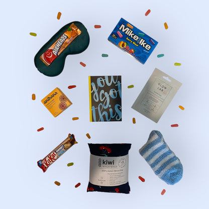 Contents of self care gift box including, silk eye mask, american candy, anihana shower steamer, you got this gratitude journal, glow lab face mask, kit kat chunky bar, kiwi wheatbag and fluffy socks.