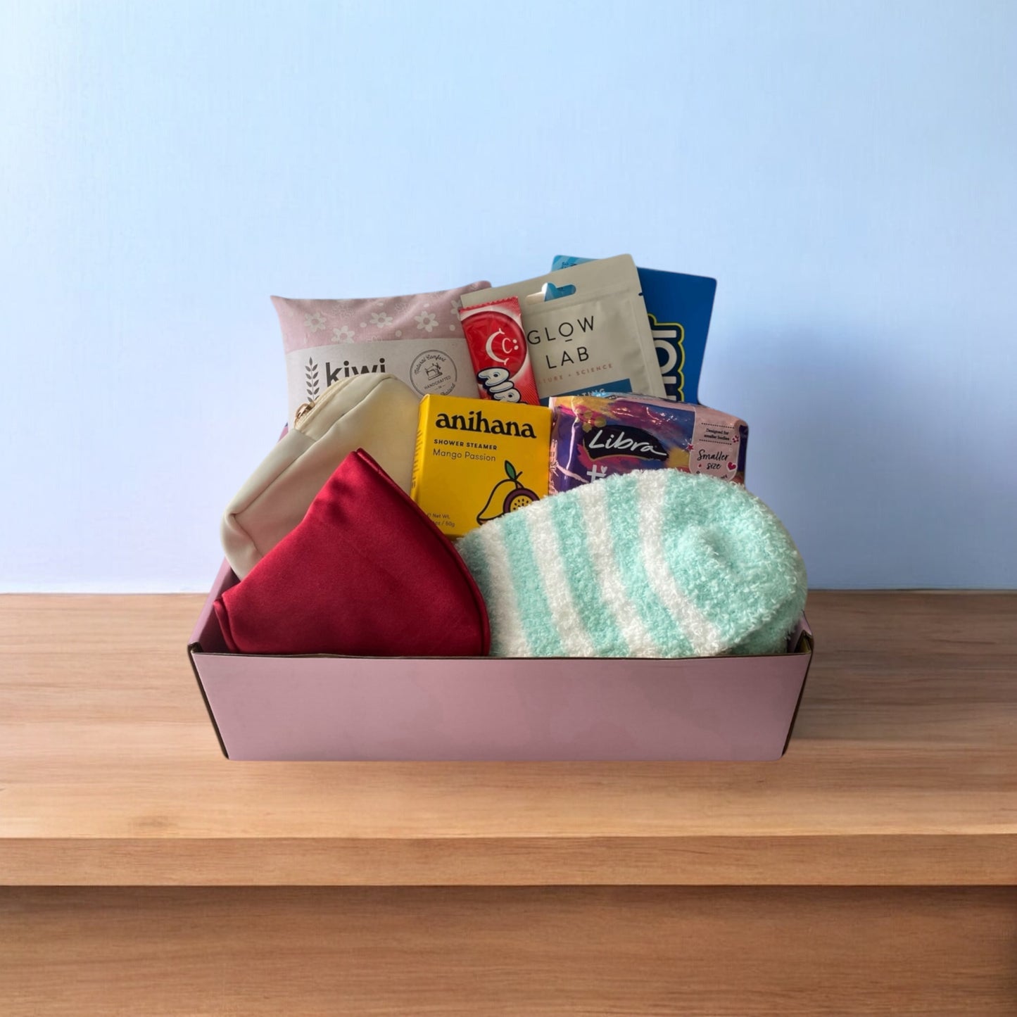 Contents of gift box displayed in gift box. Kiwi wheat bag, box of american candy,glow lab face mask, airheads candy, little purse, anihana shower steamer, libra teen pads, eye mask nd fluffy socks.