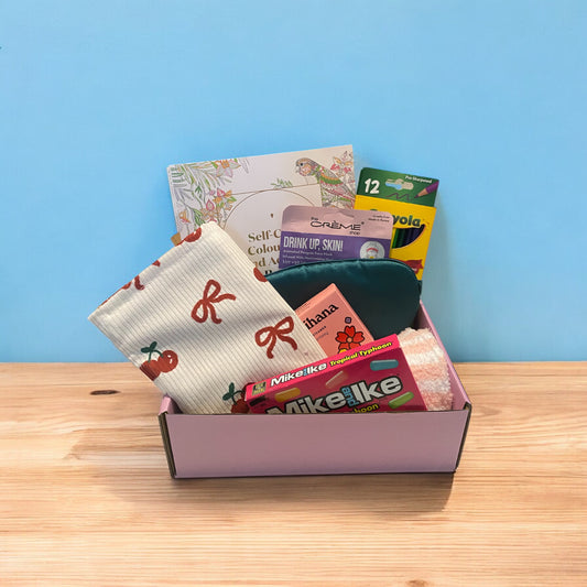 Beautiful gift box displaying items. Self care colouring books, crayola colouring pencils, face mask, eye mask, shower steamer, mike and ike candy, fluffy socks and toilet bag.