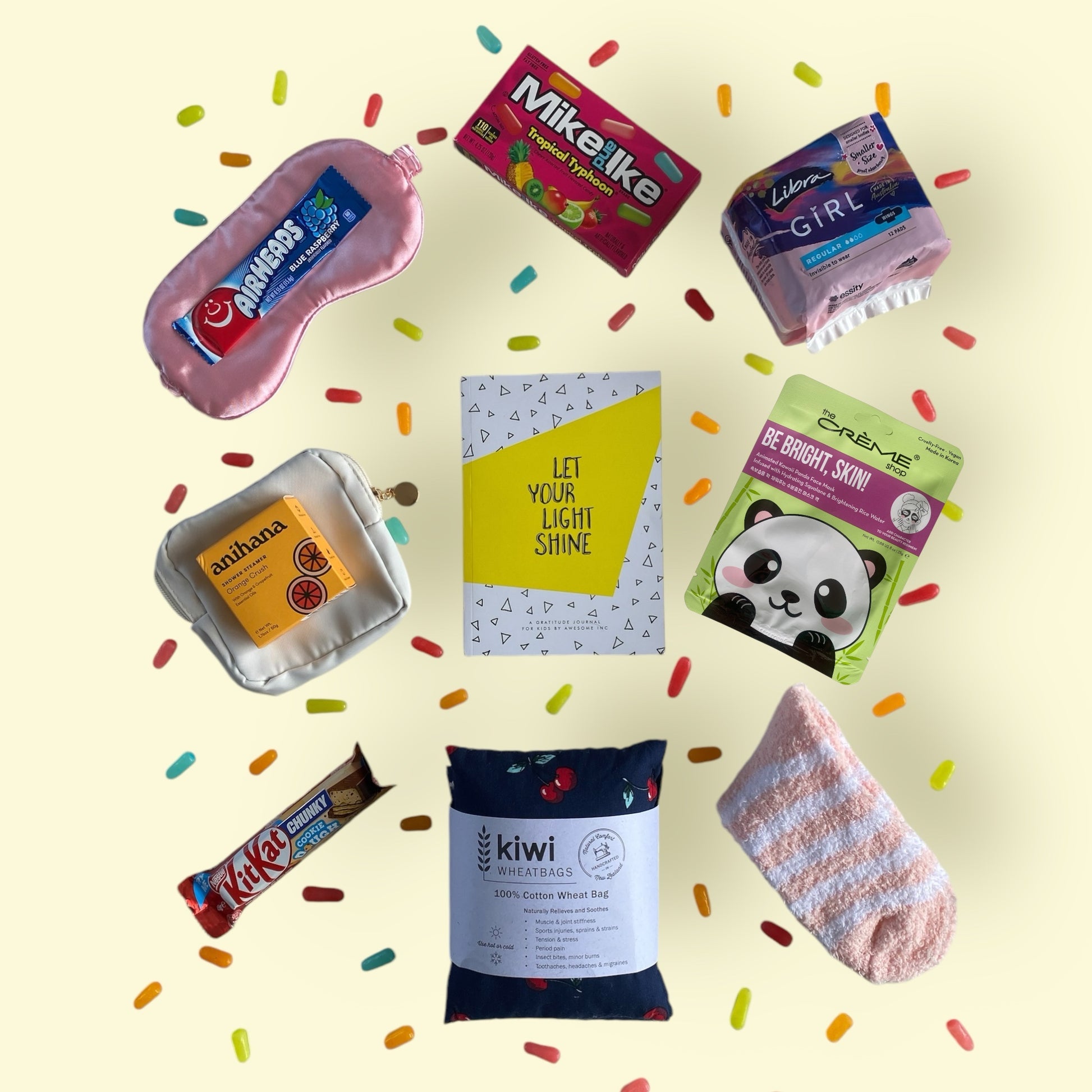 Contents of gift box displayed in the gift box. Kiwi wheat bag, gratitiude journal,glow lab face mask, Mike and Ike candy, kit kat, little purse, airhead candy, eye mask, libra teen pads, fluffy socks and anihana shower steamer.