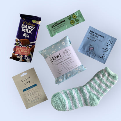 Contents of our self care gift box including, cadbury dairy milk, anihana bath salts, soleiz self warming eye mask, kiwi wheat bag, glow lab face mask and fluffy socks.